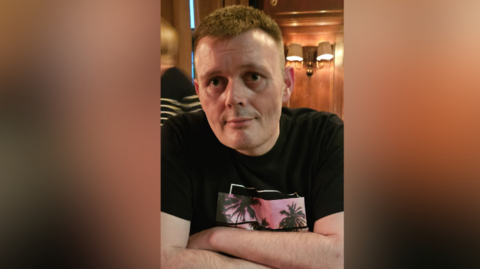 A man is sitting in a restaurant. He's wearing a black top with a design showing palm trees against a sunset. He has brown hair. A woman is passing in the background. Some light fixtures are on the wall. 