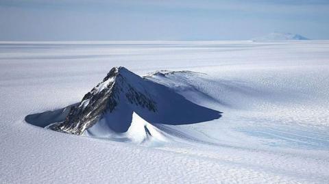 A section of the West Antarctic Ice Sheet with mountain peaks poking out of the pristine, flat, white icy surface. The sky above is light blue with light clouds.