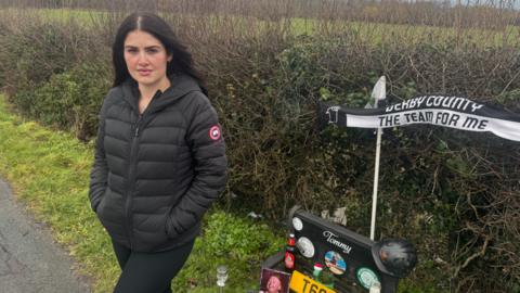 Chloe Mefford (dark hair, black jacket) standing on a roadside pavement in front of a hedge and next to a memorial to Tommy Hunter