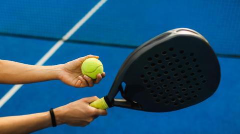 A Padel ball is held in one hand and a black Padel racket in the other. The court is blue with white lines and a black net. The person has a brown band around their right wrist. 