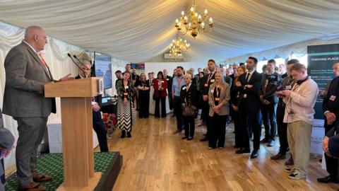 A man in a grey suit speaks to an audience of about 50 people from a lectern. They are all inside a pavilion with two chandeliers hanging from the ceiling.