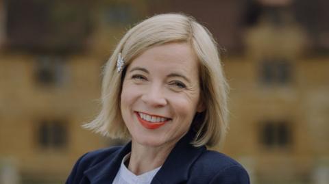 Lucy Worsley standing outside Ashby Manor, St Ledgers. She is wearing a dark coat, light shirt, is smiling and looking at the camera. She is standing in front of a historic house, with bricks and a large number of windows, the house behind is blurred. Lucy has blond chin length hair, with a side parting, and a clip to the left.