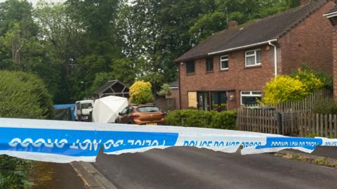 Blue police tape is seen in front of a house with boarded up windows and a white forensics tent.