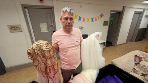 Lee Nicholson, a man with greying hair wearing a pink t-shirt and green trousers, is holding up ornate dresses while standing in what looks like a community hall. He is wearing a glum expression. 