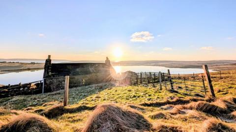 blue sky with the sun rising in the centre with a tarn, cottage and fields in the foreground 