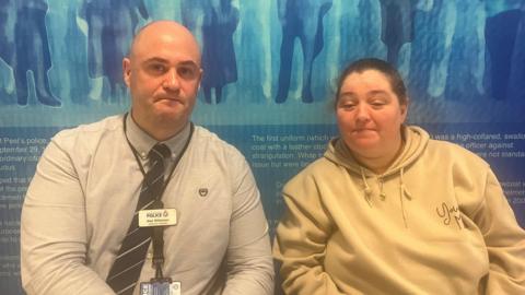 Detective Inspector Alex Wilkinson and Sarah Hayden sit together in front of a blue GMP display board. DI Wilkinson is wearing a shirt and a name tag, while Ms Hayden is wearing a cream-coloured hoodie. 