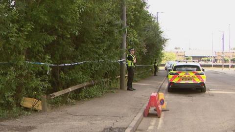 Wide shot of two police officers standing on a pavement in front of a wooded area that has been cordoned off with blue and white police tape. There is a police car among vehicles parked on the road in front of them.