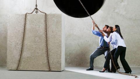 Three people straining against a pulley attached to a large white breezeblock