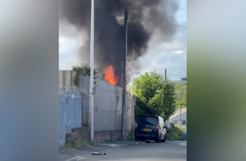 A street with a fence running along the pavement. Thick black smoke is rising up from the other side of the fence, and a large flame is visible over the top. A black car is parked nearby.