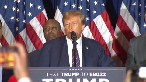 Donald Trump speaking into a microphone in front of a man and the US flag