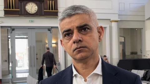 Image of the Mayor of London Sir Sadiq Khan wearing a blue suit jacket and white shirt, standing in a white panelled hall. Rows or grey chairs and a dark wood and gold clock is visible in the background