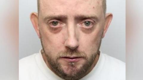 Mugshot of a mostly bald man. He looks sad and like he may have been crying, with dark circles under his eyes. He has a short light-brown beard and wears a white jumper.