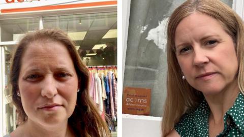 A selfie of two women standing outside a charity shop. The woman on the right has long hair and is wearing a green top with white dots. The woman on the left also has long hair and is wearing a white top. The entrance to an Acorns charity shop is behind them with the doors open and a clothes rack inside with bunting on the ceiling.