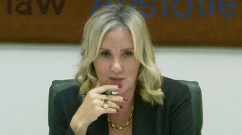 Dame Caroline Dinenage chairs a meeting in parliament. She has wavy blonde hair and wears a dark jacket and a gold chain necklace.