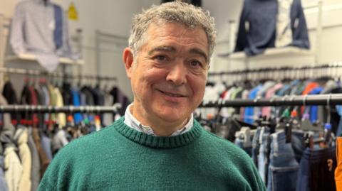 Rob Jones has short grey hair and is wearing a green jumper. He is standing in front of a clothing rack.