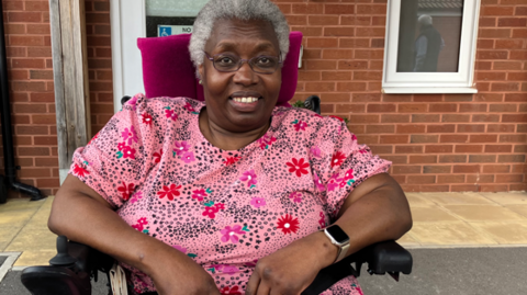 A picture of a lady sat in a wheelchair smiling at the camera. She has grey hair and is wearing glasses. She is wearing a watch on her left wrist. She is sat in front of a brick house.