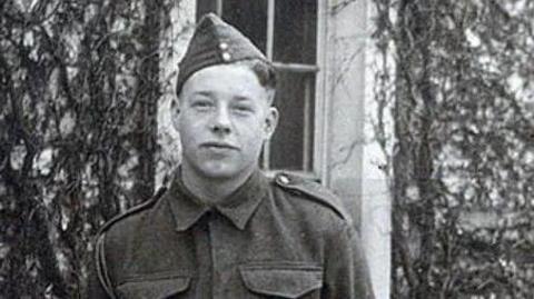 A black and white photograph of James Conway wearing military uniform and hat. He is stood in front of a white building, which has plants growing up the walls and is looking at the camera.