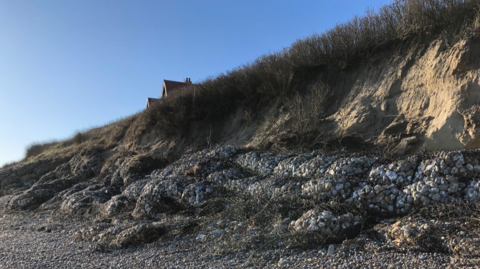 Geobags of rocks form sea defences at the foot of an eroded cliff on a gravel beach, with a house seen in the background behind long grass at the cliff edge.