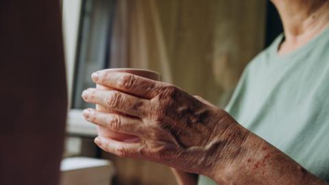 An elderly person holding a mug while looking out of a window.