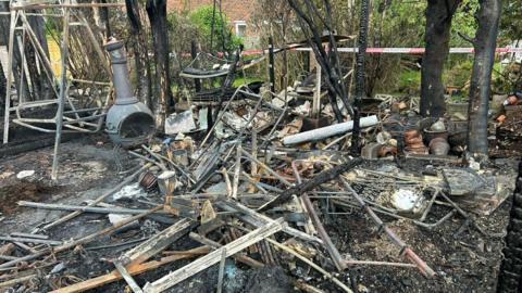 A pile of burned wreckage sat on a charred surface in a garden. The remains of some trees, a garden swing and a garden stove can be seen amongst the ruins.
