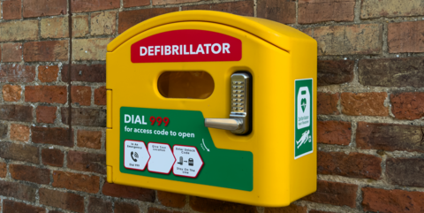 A yellow defribulator box mounted on a brick wall with the word "Defibrillator" in red on it, along with instruction diagrams on its use