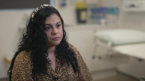 A woman wearing a leopard-print shirt sat in a doctor's room. A white doctor's bed is in the background. She is sat down, looking past the camera and out into the distance.