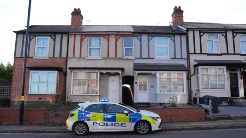 A police car parked outside the penultimate house at the end of a row of terraced houses. The red-brick and rendered houses have two stories and timbers painted in black, red and blue.