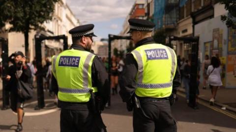 Image of two uniformed police officers stood with their backs to the cameras as people enter gates to Notting Hill Carnival in 2024