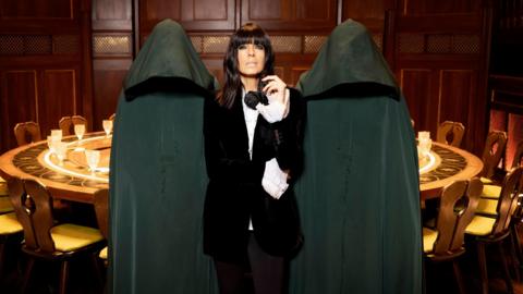 Presenter Claudia Winkleman is dressed in black and flanked by two hooded figures. They are standing in front of a large, ornate round table.