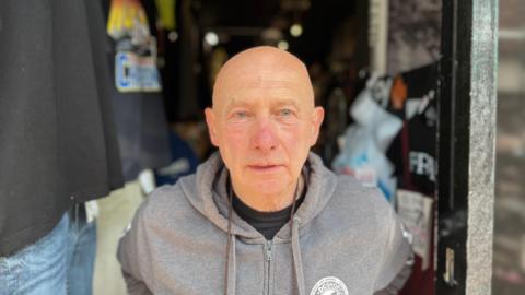 A bald, white man wearing a grey hooded jumper. He is stood in the doorway of a shop.