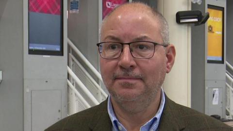 A man wearing a suit and shirt standing in front of check-in desks at Jersey Airport.
