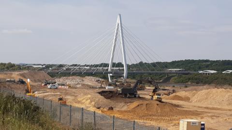 The construction site of the Crown Works film studio. A large area of land is dug out. There are diggers dotted around the site. A white bridge borders is on one side of the site.