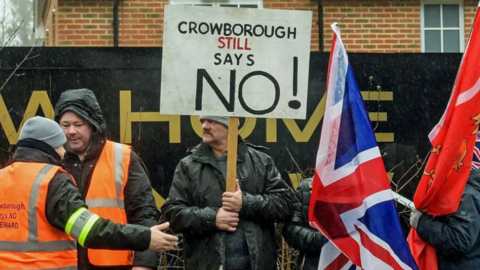 A man in a black jacket is holding a sign saying Crowborough Still Says No, with two people in orange hi vis vests on his left, and people carrying flags to his right