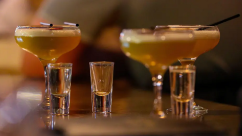 Three martini glasses with short black straws on top of them, containing a cocktail, along with three shot glasses, all on a table.