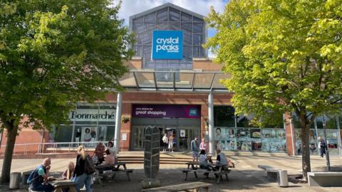 Outside the doors of a shopping centre. Shoppers sit in the sunshine on benches.