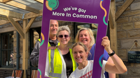 Four smiling people standing close together outdoors, posing for a photograph using a large rectangular purple frame held in front of them.