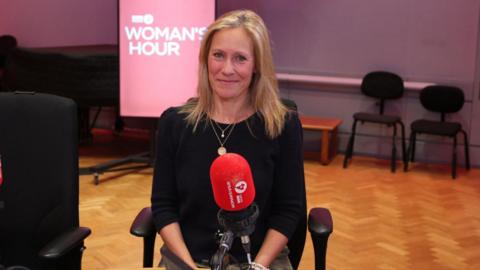 Sophie Raworth smiling, sat in a chair in a radio studio with a red mic in front of her.