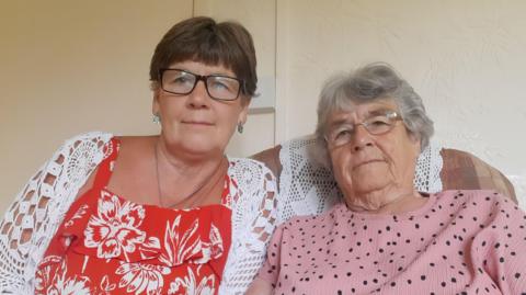Karen Williamson is wearing glasses and a red flowery top beneath a light white cardigan. Jean Hewitt, who also wears glasses, has a pink, black spotted jumper on. They're sitting together on an armchair.