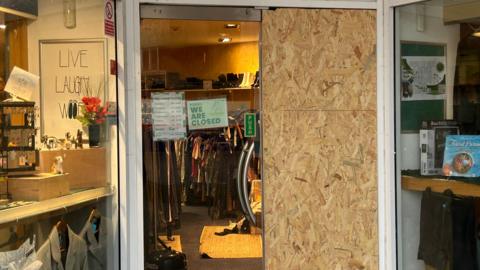 The entrance to the Oakwood Dog Rescue charity shop in Hull. The right hand door is boarded up having been smashed. The left hand door has a "we are closed" sign on it. You can see through the door to the inside of the shop.