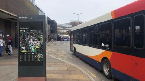 A white, red and blue bus puling in to a stop. On the pavement is a sign that reads Bus Station above a map of Guildford.