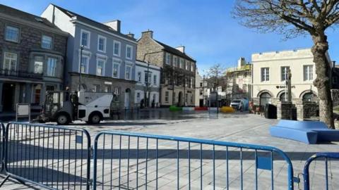 Market Square, a large area with grey tiling, there are stone buildings in the background, and a blue barrier in the front, there is a street cleaner vehicle, and no othervehicles in the space.