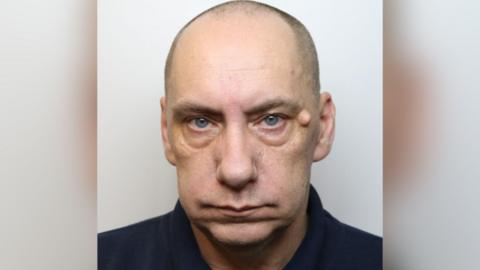 A custody shot of Mark Bellas. He is bald and has blue eyes. He is pictured against a white background, looking straight at the camera with a blank expression.