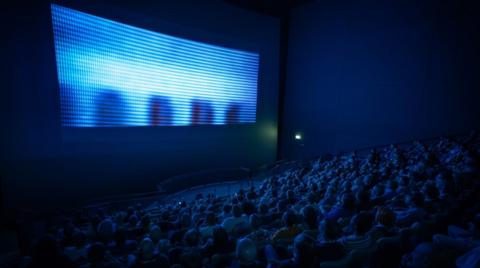 A full cinema with people watching a large screen but the projection doesn't fill up the whole screen.