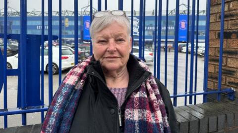 A woman standing in front of blue railings outside Birmingham City Football Club. She is wearing a black coat with a blue and purple checked scarf over it. She has short grey hair and is looking sad.