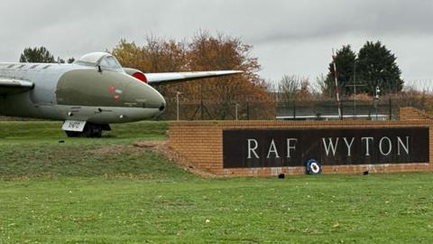 An old-looking jet plane outside RAF Wyton. The plane is grey and khaki green, and is sat on top of a raised, grassy platform. In front of its nose cone, on the right, is a name plate bearing "RAF Wyton", set into a low brick wall. There is an expanse of grass in the foreground and grey skies above the plane. In the background are autumnal-looking trees, whose leaves have turned orange.