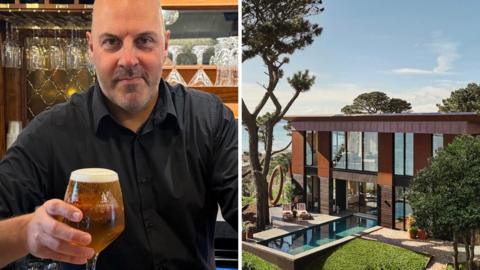 Darren Summers holds up a pint of beer while standing behind a pub. Cornish house