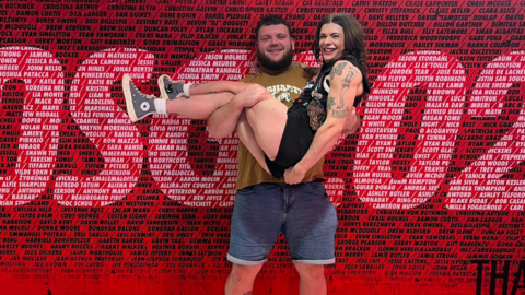 Joe is holding Rosie in his arms with a bright red poster backdrop behind them. He is wearing shorts and a t'shirt and she is in shorts and a black vest top.