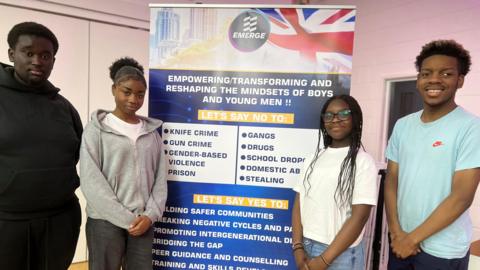 A group of four young people stand in front of a sign promoting the Emerge project, which pledges to "reshape mindsets".