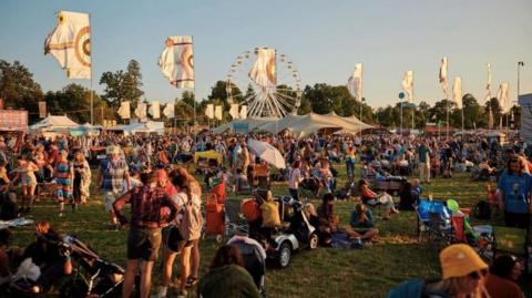 A scene of people gathered at Wiltshire's Womad festival in 2024. Crowds are sitting and standing on the grassy site as colourful flags catch the wind, bathed in evening sunlight.