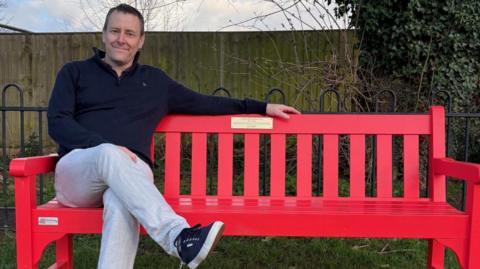 Nick Thomas, wearing a blue jumper and grey trousers, has short brown hair. He is sitting on a bright red bench.